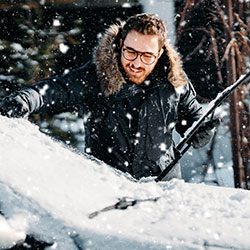 A man brushing the snow off his car while it flurries around him.