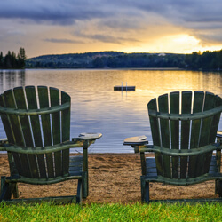 Two Muskoka chairs on a beach looking out over the water at sunset.