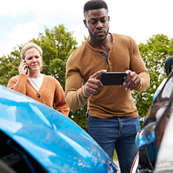 A man taking a photo of the damages caused from being rear-ended by another driver who is on the phone calling her insurer.